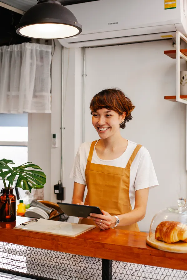 Small business owner learning how to start using AI in small business, checking a tablet at her shop counter while an AI agent handles admin tasks
